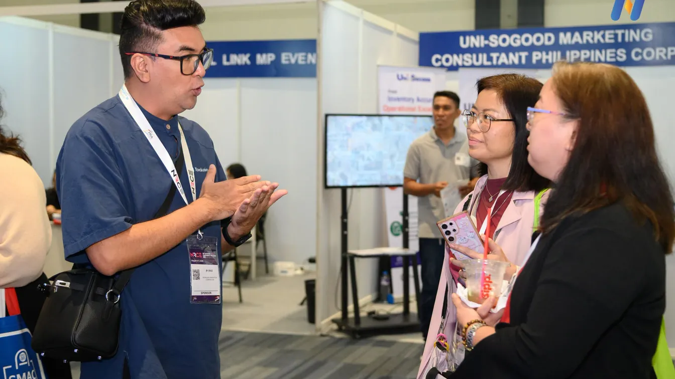 Novaluxia booth at a trade show, an exhibitor explaining the platform to two visitors, one of the public appearances our "Is Novaluxia Legit" check relies on